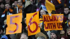 Football supporters hold up claret-and-amber paper signs bearing the words
