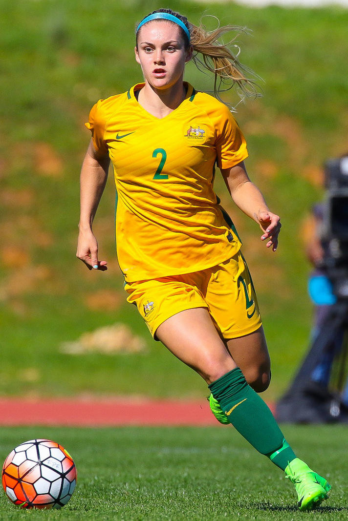 A person in a yellow and green team uniform runs toward a ball on a football pitch. 