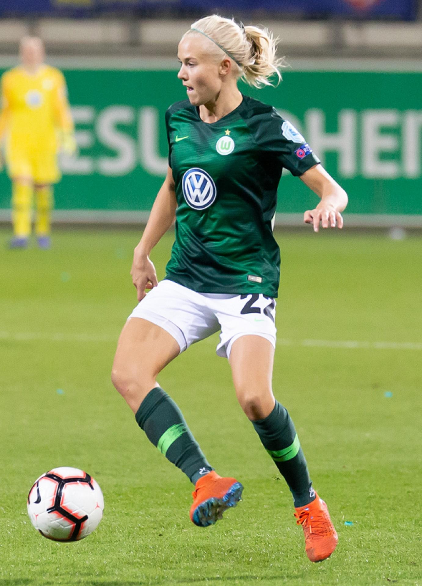 A person wearing a green sports jersey and white sports on a football pitch, next to a ball. 