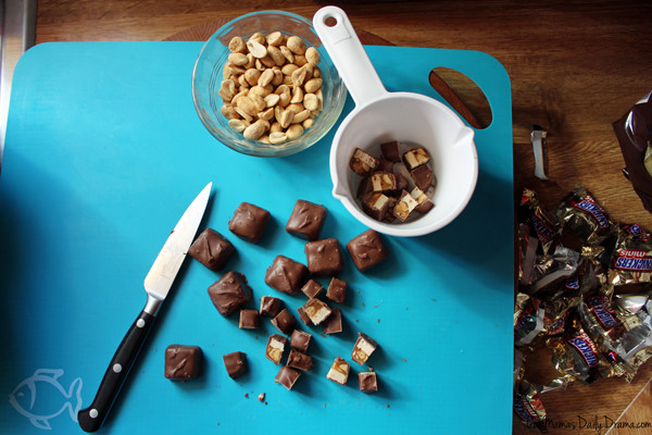 a knife on a blue cutting board with a bowl of peanuts and a handful of mini snickers bars