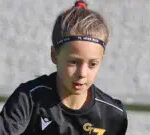 Poppy plays football. She has dark hair pulled back into a ponytail and is wearing a black football kit. She is looking down at the ground