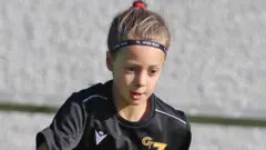 Poppy plays football. She has dark hair pulled back into a ponytail and is wearing a black football kit. She is looking down at the ground