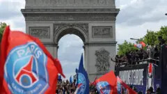 PSG parading down towards the Arc de Triomphe on an open-top bus, fans holding PSG flags