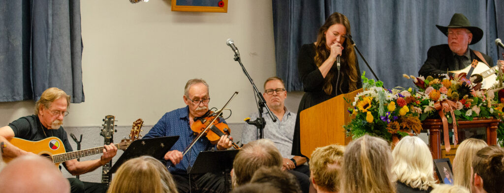Musicians John Park Wheeler, Brian Hebert and Clint Degarie accompany Bailey Rogers as she sings A Way Down the River at the memorial service for Steve Agnew.