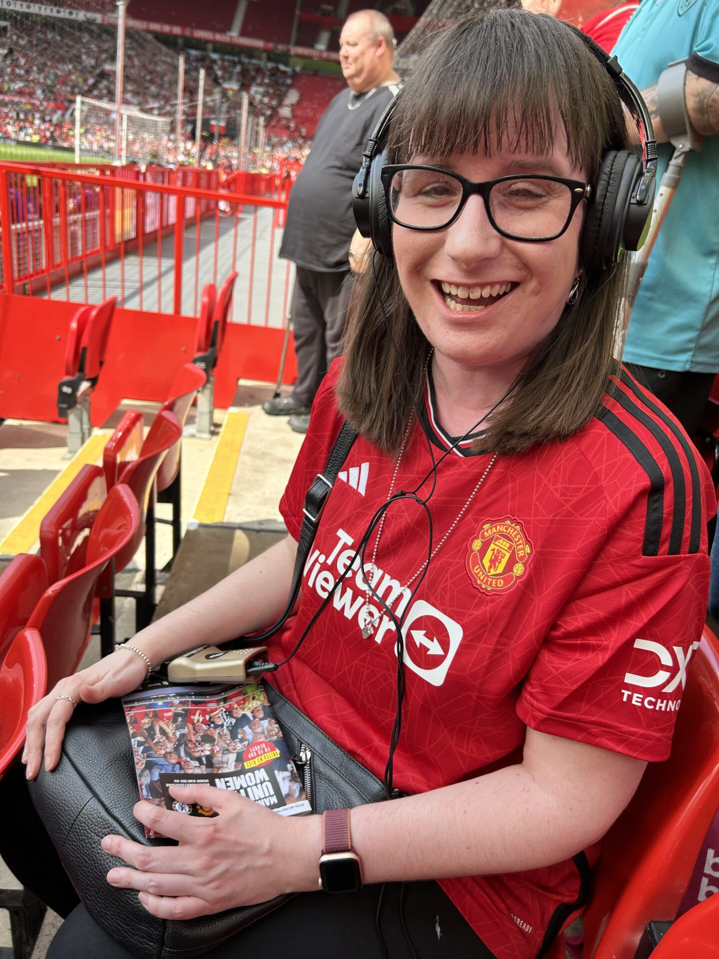 Holly sat in the stadium with the Manchester United match programme and headset controls resting on her knee. She has a headset on and is wearing a Manchester United shirt.